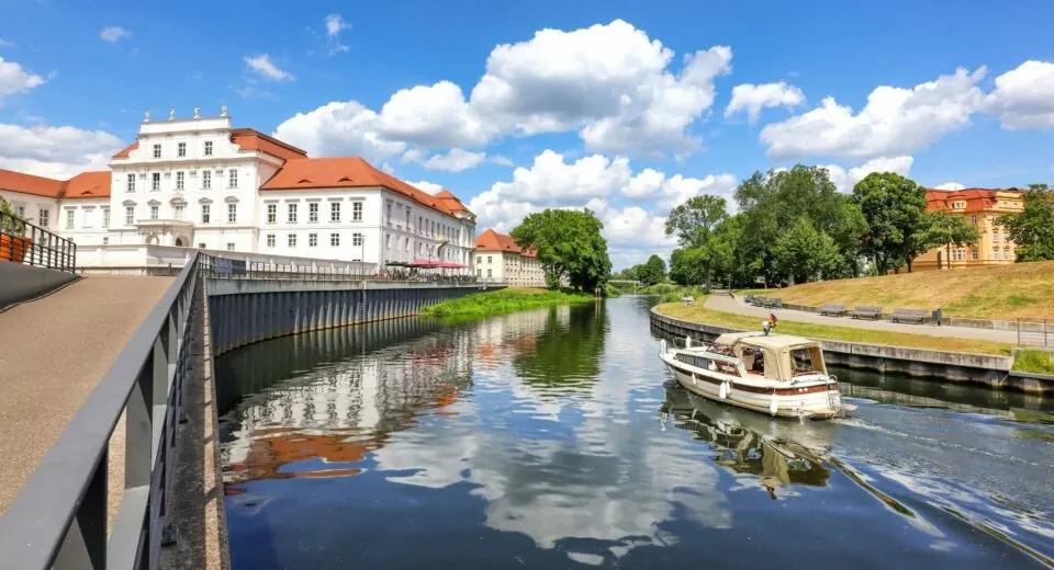 Eine ruhige Kanalszene in Oranienburg zeigt ein weißes Boot, das auf dem ruhigen Wasser kreuzt. Auf der linken Seite ragt ein weißes historisches Gebäude mit rotem Dach hervor, während die rechte Seite von üppigem Grün und weiteren Gebäuden gesäumt ist. Der Himmel ist strahlend blau mit vereinzelten Wolken.