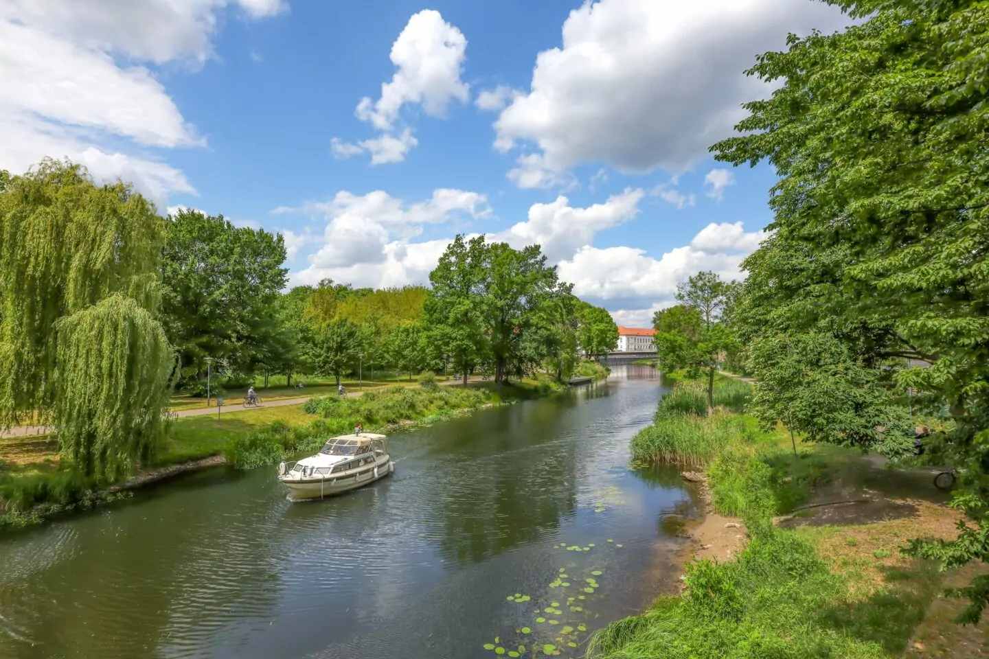 Eine ruhige Flussszene in Oranienburg mit einem kleinen weißen Boot, das auf dem ruhigen Wasser kreuzt, umgeben von üppigen grünen Bäumen und Pflanzen unter einem teilweise bewölkten, blauen Himmel. In der Ferne ist schwach eine Brücke zu erkennen, flankiert von weiterem Grün.