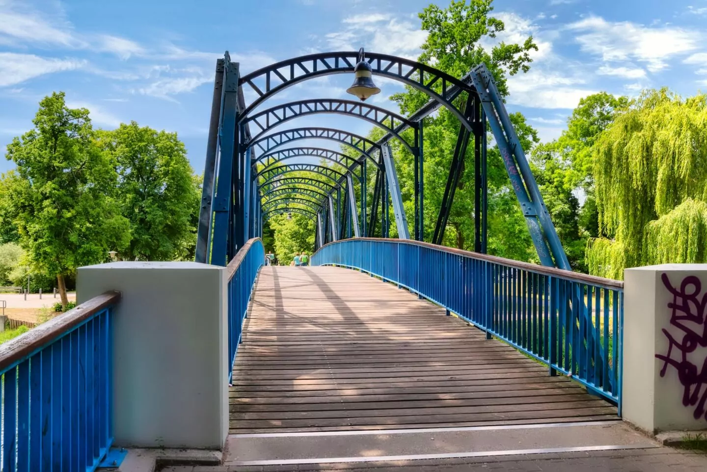 Eine blaue Fußgängerbrücke aus Metall und Holz mit geschwungenen Balken, umgeben von üppigen grünen Bäumen an einem klaren, sonnigen Tag in Oranienburg. Auf dem rechten vorderen Pfeiler der Brücke ist ein kleines Graffiti zu sehen. Die Brücke führt in eine Parkanlage.