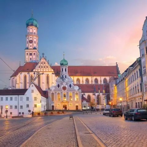 Eine malerische Ansicht von Augsburg, Deutschland, mit Kopfsteinpflasterstraßen und historischer Architektur. Das Bild zeigt die Abtei St. Ulrich und Afra bei Sonnenuntergang mit ihrer kunstvollen Fassade und den Zwillingstürmen. Die umliegenden Augsburger Gebäude und Straßenlaternen tragen zur ruhigen Atmosphäre bei.