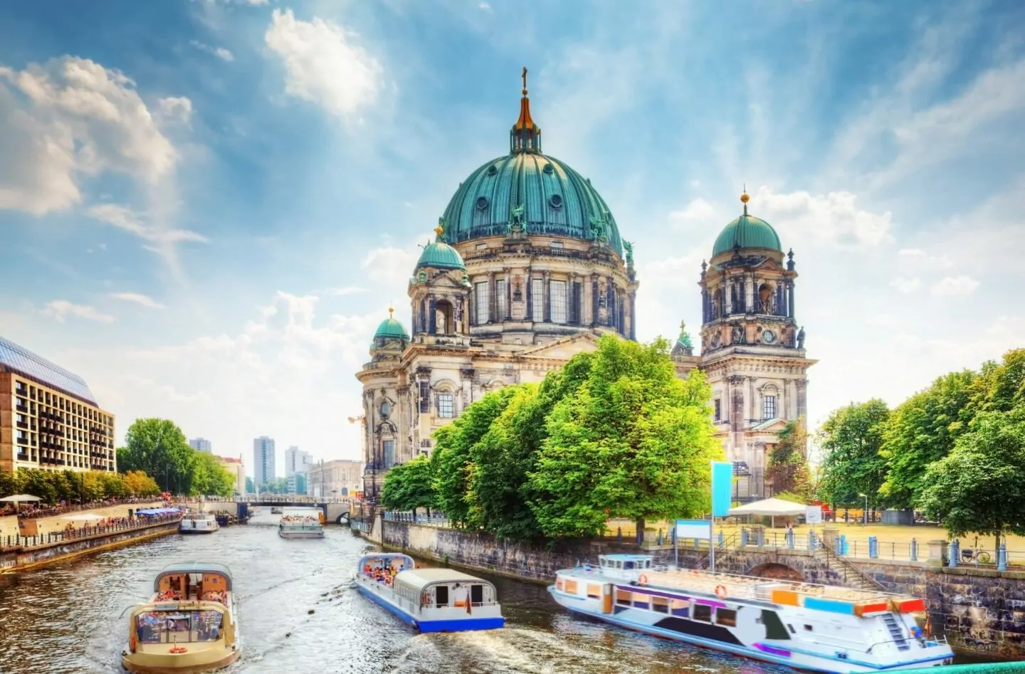 Ein Blick auf den Berliner Dom mit seinen grünen Kuppeln an einem sonnigen Tag. Im Vordergrund sieht man mehrere Boote, die auf dem Fluss fahren, gesäumt von Bäumen und historischen Berliner Gebäuden. Ein klarer blauer Himmel mit vereinzelten Wolken verstärkt die lebendige Szene.