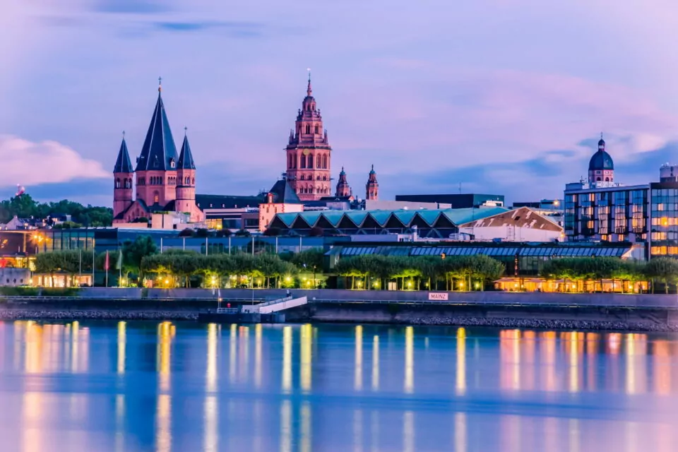 Ein ruhiger Blick auf Mainz in der Abenddämmerung zeigt seine historische Architektur. Hohe Kirchtürme und eine markante Kathedrale heben sich von einem schwach beleuchteten Himmel ab. Der ruhige Fluss im Vordergrund spiegelt die beleuchteten Gebäude und trägt zur malerischen Szene bei.