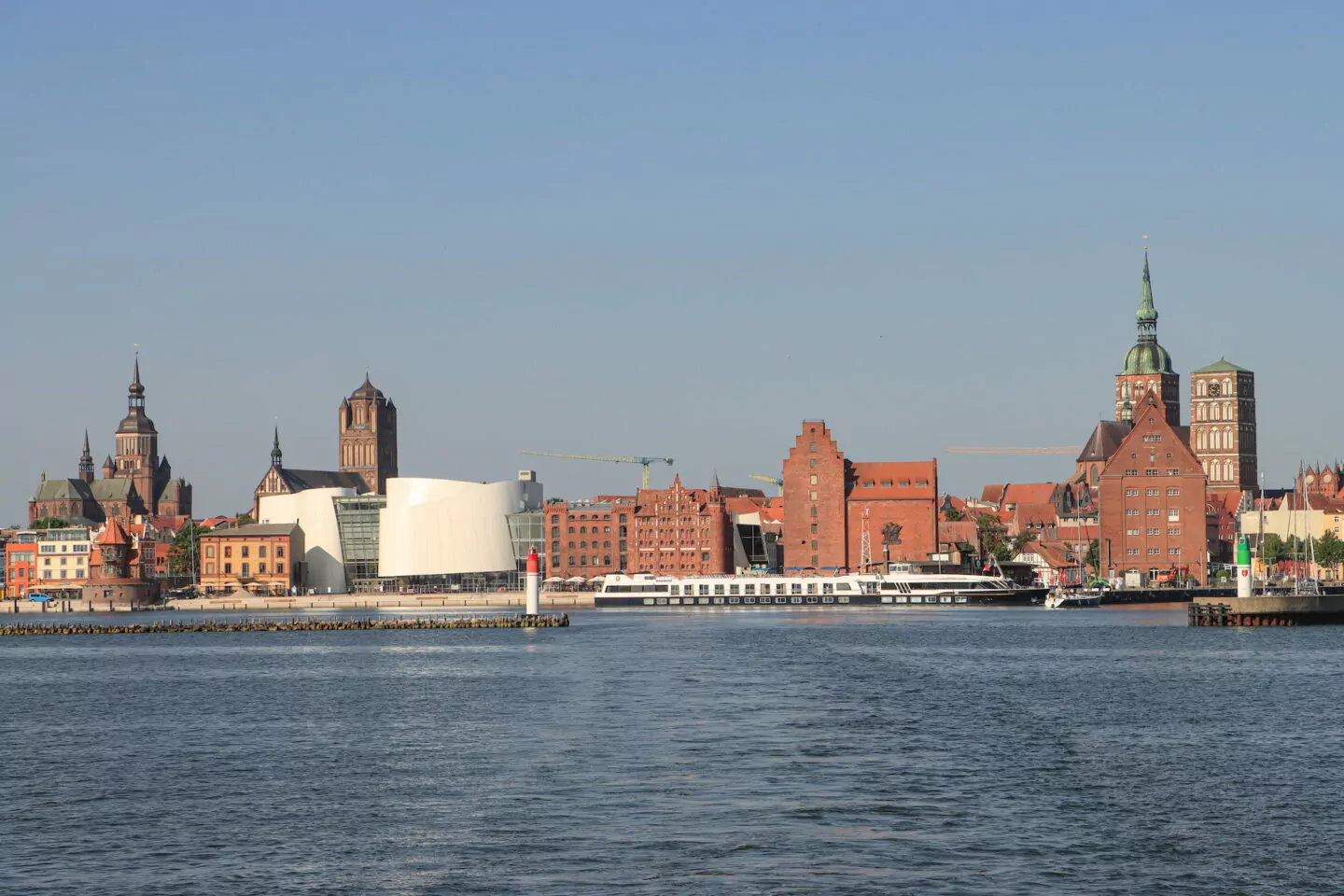 Ein malerischer Blick auf die Uferpromenade der historischen Stadt Stralsund mit markanten Backsteingebäuden und roten Dächern, Kirchtürmen und einem weißen modernen Gebäude. Ein Boot liegt am Ufer und im Vordergrund erstreckt sich das ruhige Wasser unter einem klaren blauen Himmel.