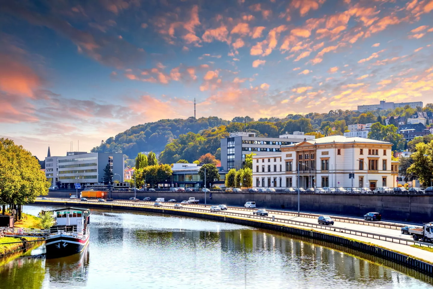 Ein ruhiger Blick auf das Flussufer in Saarbrücken bei Sonnenuntergang, der einen Kanal mit einem angedockten Boot und nahe gelegenen Gebäuden zeigt. Im Hintergrund ist ein üppig bewaldeter Hügel mit einem Turm und einer befestigten Struktur zu sehen. Der Himmel ist lebhaft mit vereinzelten Wolken, die warme Farbtöne reflektieren.
