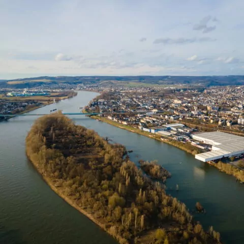 Luftaufnahme eines großen Flusses, der sich durch die dicht besiedelte Stadt Neuwied schlängelt. Im Fluss liegt eine mit Bäumen bewachsene Insel, während Wohnhäuser, Lagerhäuser und Hügel mit vereinzelten Grünflächen das Gebiet unter einem teilweise bewölkten Himmel umgeben.
