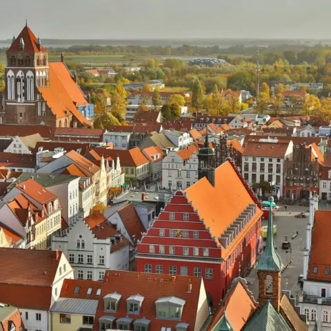 Luftaufnahme der europäischen Stadt Greifswald mit Gebäuden mit roten Dächern und einer markanten Kirche mit hohem Turm. Die Stadt ist von grünen Feldern und Bäumen umgeben und verbindet moderne und historische Architektur. Das Herbstlaub verleiht der Landschaft eine lebendige Farbpalette.