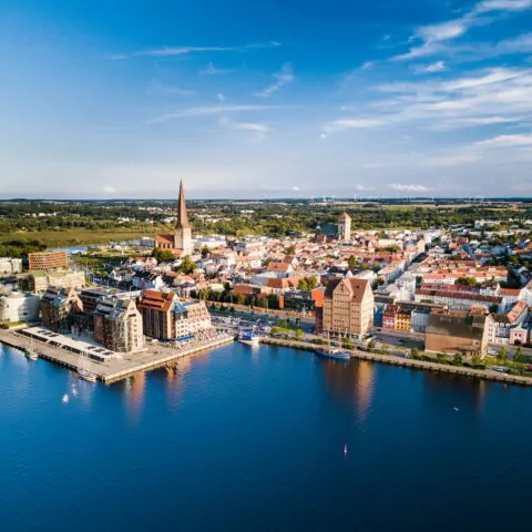 Luftaufnahme der Küstenstadt Rostock mit historischen Gebäuden und Kirchen entlang der Uferpromenade. Das Stadtbild umfasst einen markanten Turm und moderne Gebäude, die von Wasser umgeben sind, vor einer grünen Kulisse und einem teilweise bewölkten blauen Himmel.