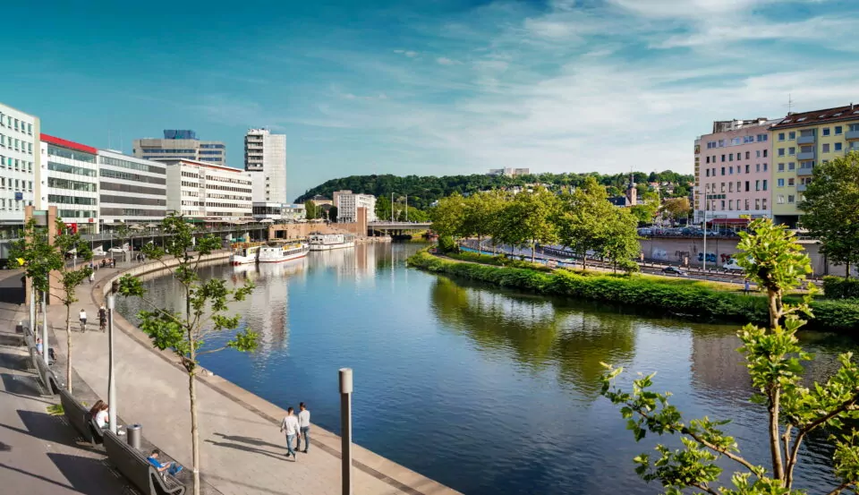 Eine malerische Stadtlandschaft am Flussufer in Saarbrücken mit modernen Gebäuden, einem Spazierweg und Bäumen am Wasser. Zwei Menschen gehen den Weg entlang, während ein Boot am Flussufer angedockt ist. Der Himmel ist klar mit ein paar Wolken und im Hintergrund ist üppiges Grün zu sehen.