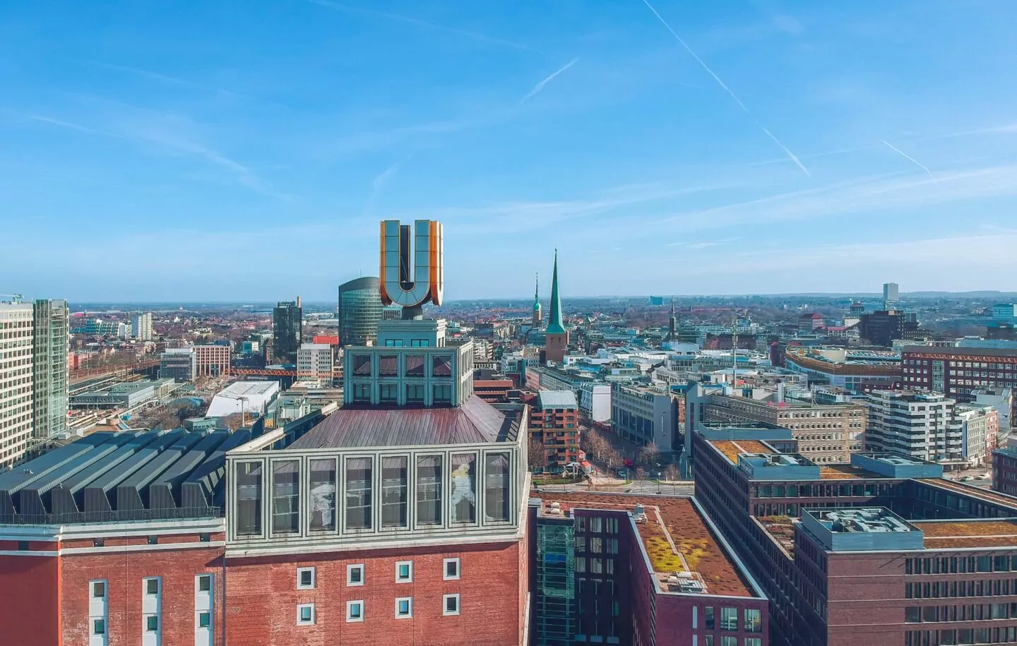 Ein Panoramablick auf die Stadtlandschaft Dortmunds an einem klaren Tag. Im Vordergrund ist ein großes, markantes rotes Backsteingebäude mit einer markanten „U“-Skulptur zu sehen. Die Stadt ist dicht mit verschiedenen Gebäuden bebaut und in der Mitte ist ein Kirchturm zu sehen.