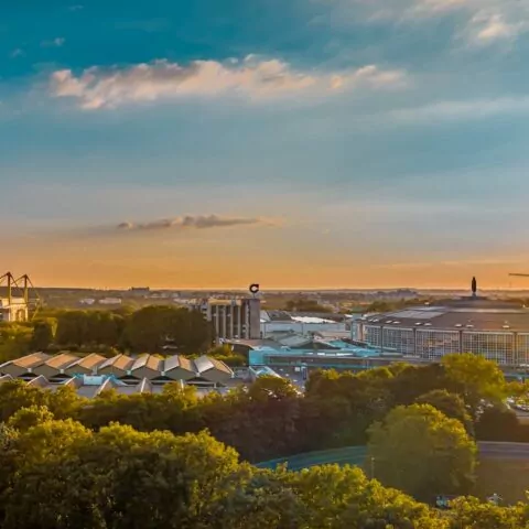 Ein Panoramablick auf Dortmund bei Sonnenuntergang zeigt eine Mischung aus moderner und traditioneller Architektur. Die Skyline umfasst ein Stadion mit Kränen im Hintergrund. Im Vordergrund ist eine üppige Grünfläche mit Bäumen zu sehen, die der städtischen Umgebung dieser deutschen Stadt natürliche Schönheit verleiht – ein Genuss für Reisende.