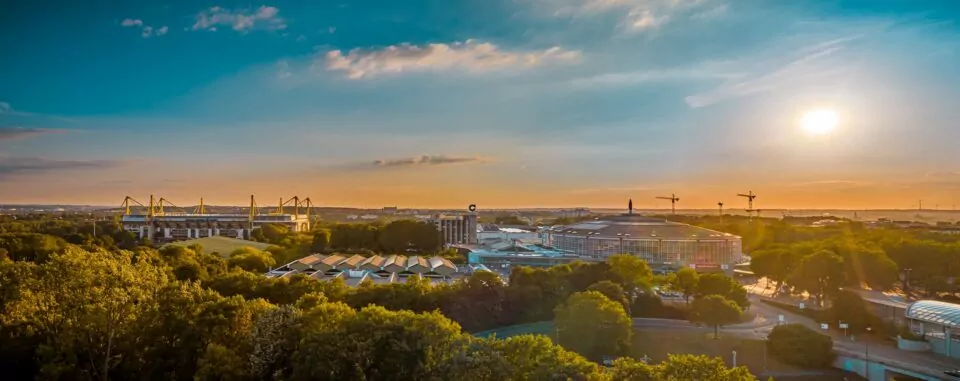 Ein Panoramablick auf Dortmund bei Sonnenuntergang zeigt eine Mischung aus moderner und traditioneller Architektur. Die Skyline umfasst ein Stadion mit Kränen im Hintergrund. Im Vordergrund ist eine üppige Grünfläche mit Bäumen zu sehen, die der städtischen Umgebung dieser deutschen Stadt natürliche Schönheit verleiht – ein Genuss für Reisende.