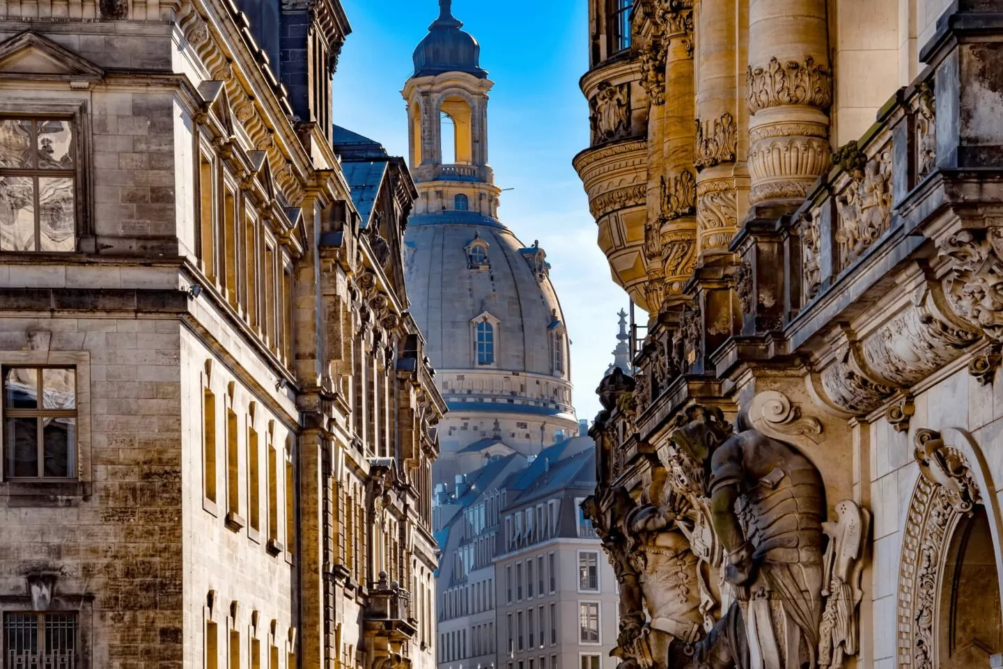 Eine schmale Straße in einer alten europäischen Stadt, wahrscheinlich Dresden in Sachsen, mit historischer Architektur und dekorativen Steinmetzarbeiten an den Gebäuden entlang der Straße. Im Hintergrund ragt eine große Kuppelkirche markant vor einem klaren blauen Himmel auf und fängt das Wesen dieser bezaubernden Ecke Deutschlands ein.