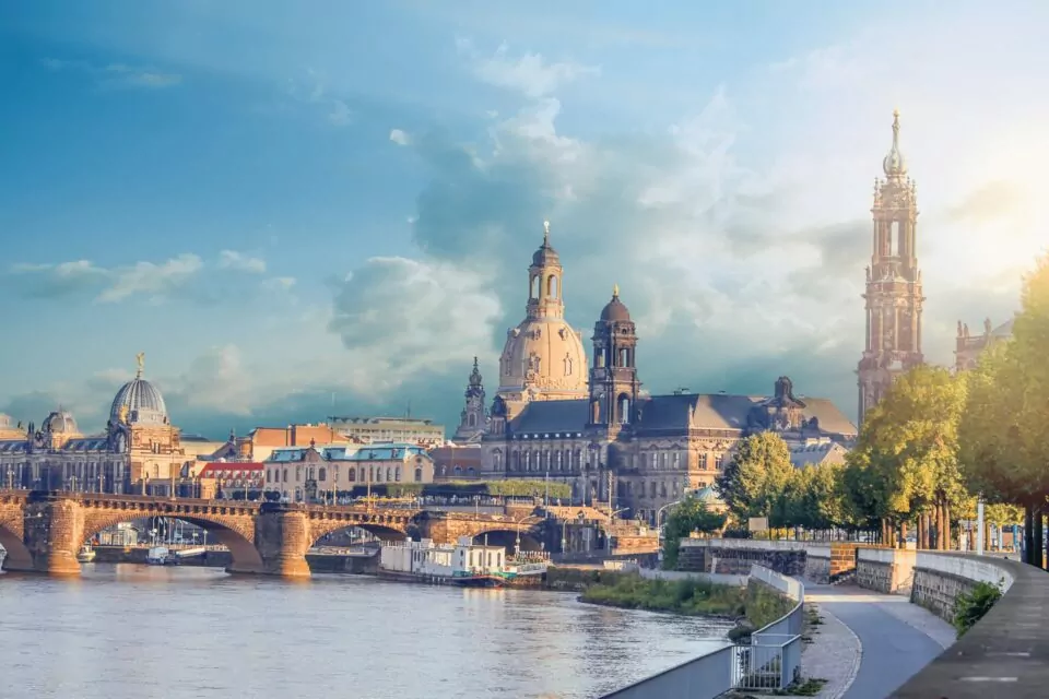 Eine malerische Aussicht auf Dresden, Deutschland. Das Bild zeigt historische Architektur entlang der Elbe, darunter die Frauenkirche und den Dresdner Dom. Der Himmel ist teilweise bewölkt und die Sonne beleuchtet die Gebäude. Bäume säumen den Uferweg – ein perfektes Ziel für Reiseliebhaber, die reiche Geschichte und Schönheit suchen.