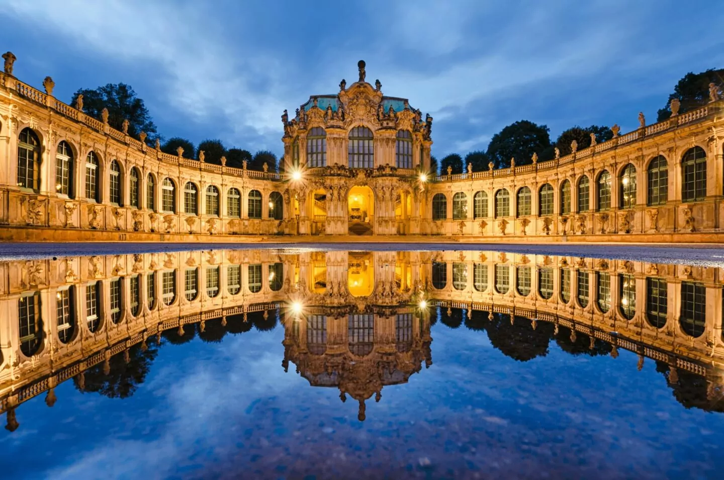 Elegantes Barockschloss in Dresden, beleuchtet in der Abenddämmerung, spiegelt sich in einem ruhigen Teich. Das Gebäude zeichnet sich durch komplizierte architektonische Details und symmetrische Kurven aus. Der Abendhimmel verstärkt den goldenen Glanz des Gebäudes und schafft eine majestätische und ruhige Szene.