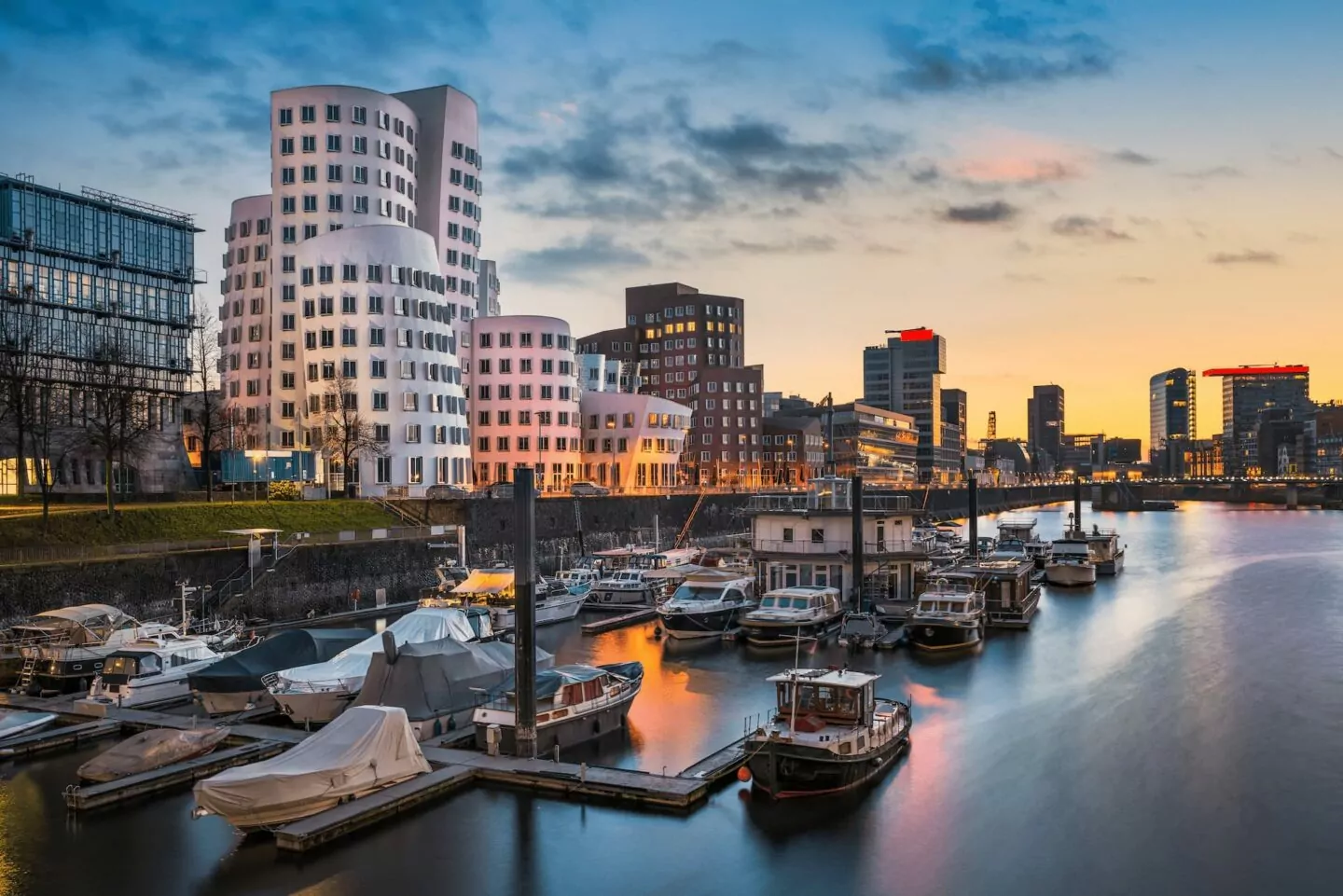 Ein ruhiger Hafen in Düsseldorf bei Abenddämmerung mit angedockten Booten, modernen Gebäuden und einzigartigen, welligen weißen Strukturen, die sich im ruhigen Wasser spiegeln. Der Himmel wechselt von Blau zu Orange, während die Sonne hinter der Skyline der Stadt untergeht.