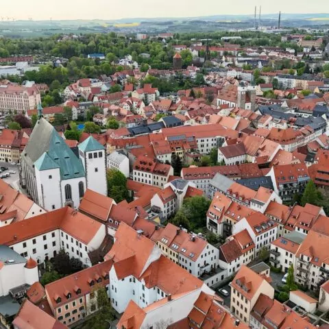 Luftaufnahme der historischen europäischen Stadt Freiberg mit roten Ziegeldächern. Auffällige Merkmale sind eine große Kirche mit zwei hohen Türmen und eine Mischung aus Wohn-, Geschäfts- und Industriegebäuden. Umgeben von Grün und fernen Hügeln unter einem klaren Himmel.