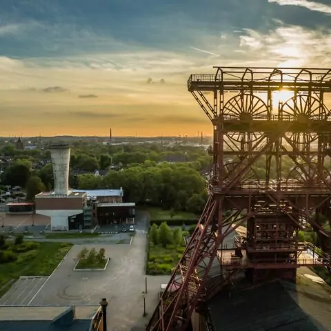 Luftaufnahme eines Industriegeländes in Gelsenkirchen mit einer großen Metallkonstruktion im Vordergrund, möglicherweise ein Fördergerüst, bei Sonnenuntergang. Gebäude und üppiges Grün umgeben das Gebiet, in der Ferne ist die Skyline der Stadt unter einem teilweise bewölkten Himmel zu sehen.