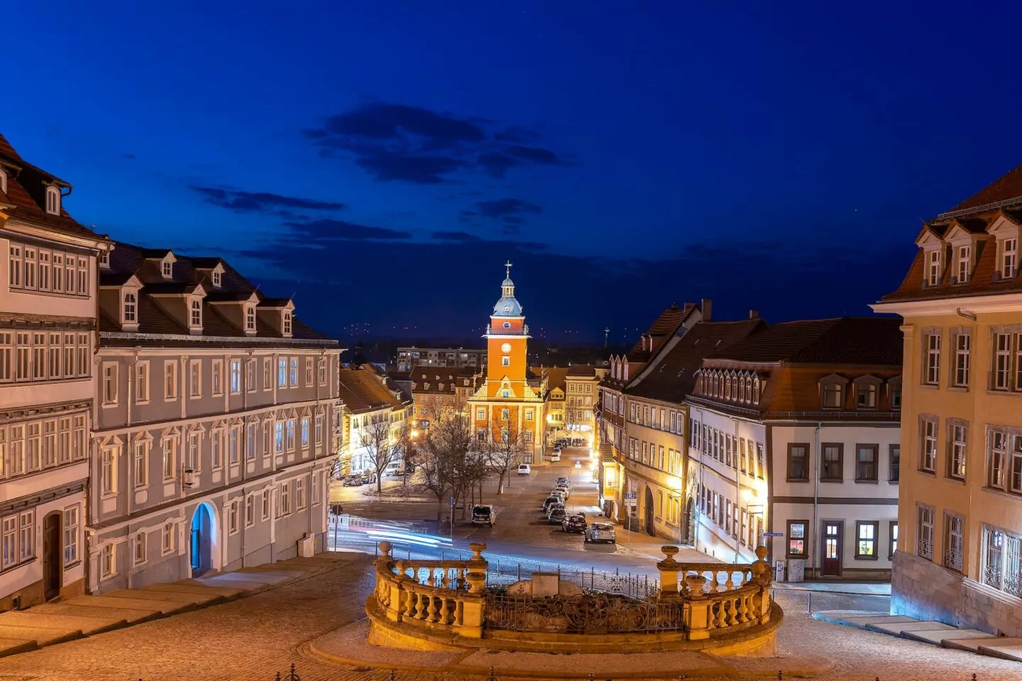 Ein nächtlicher Blick auf einen bezaubernden europäischen Stadtplatz in Gotha mit historischen Gebäuden, die von Straßenlaternen beleuchtet werden. In der Mitte sticht ein markanter Uhrturm vor dem dunkelblauen Himmel hervor. Entlang der Straßen sind Autos geparkt und die Szene ist ruhig und malerisch.