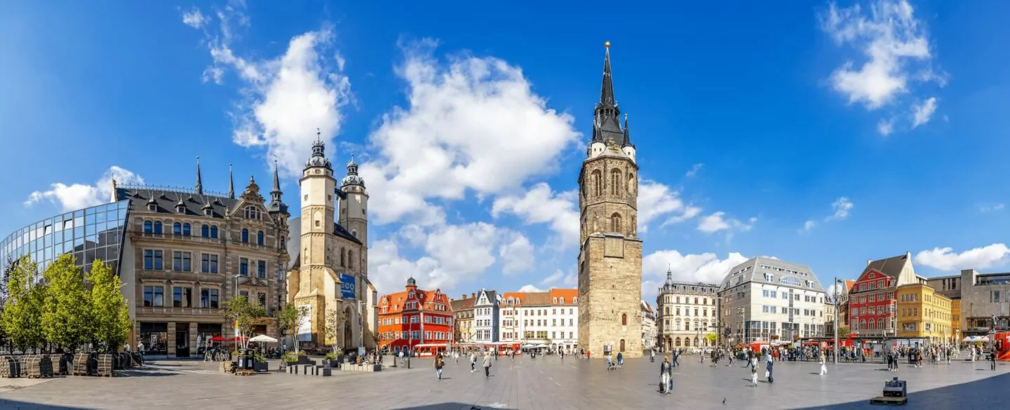 Ein Panoramablick auf den belebten Stadtplatz von Halle an einem klaren, sonnigen Tag. Der Platz ist von historischen Gebäuden mit unterschiedlicher Architektur umgeben, darunter ein hoher Kirchturm in der Mitte. Menschen laufen herum, und man sieht Cafés und Restaurants mit Sitzgelegenheiten im Freien.