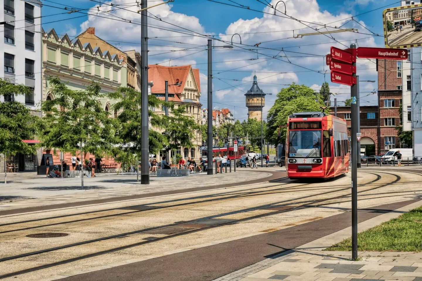 Eine lebendige Stadtszene in Halle mit einer roten Straßenbahn, die auf den Gleisen fährt. Die umliegenden Gebäude sind in verschiedenen Architekturstilen gehalten, darunter ein Turm im Hintergrund. Menschen sitzen und gehen in einer von Bäumen gesäumten Gegend umher und genießen das sonnige Wetter.