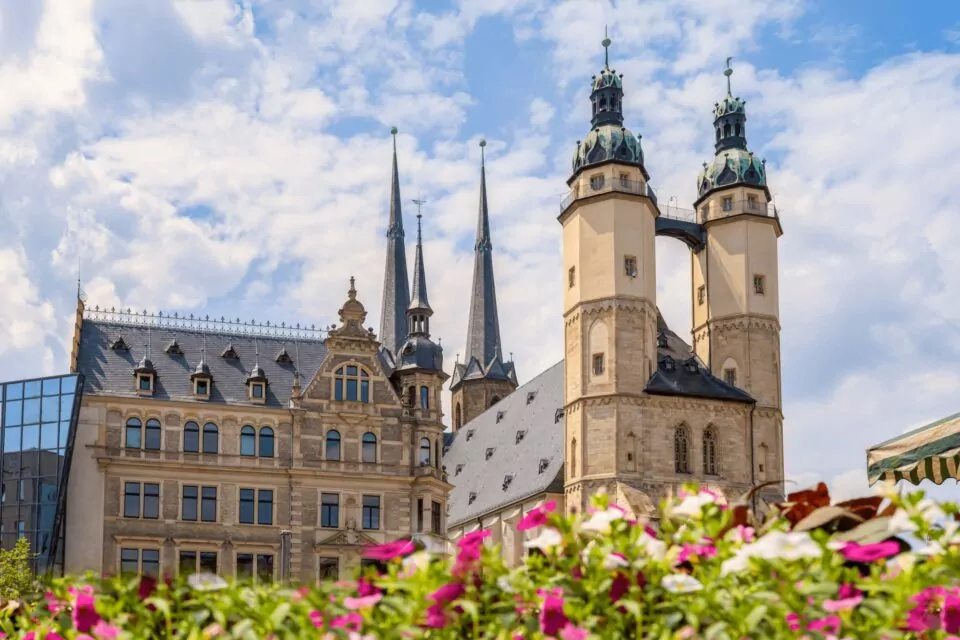 Eine europäische Stadtansicht in Halle zeigt eine markante historische Kirche mit Zwillingstürmen und mehreren Kirchtürmen. Im Vordergrund ist eine farbenfrohe Anordnung von rosa und weißen Blumen zu sehen, während der Hintergrund einen teilweise bewölkten Himmel mit einer Mischung aus alten und modernen Gebäuden zeigt.