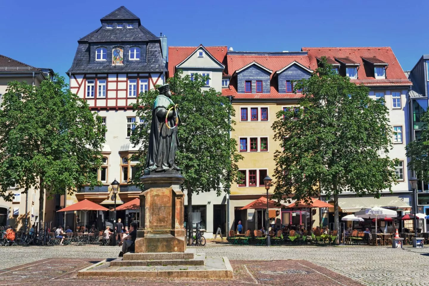 Auf einem öffentlichen Platz in Jena steht in der Mitte eine alte Statue einer vermummten Gestalt. Im Hintergrund sind eine Reihe farbenfroher Gebäude im europäischen Stil mit roten Ziegeldächern und Dachfenstern zu sehen. Man sieht Bäume und Straßencafés mit Sonnenschirmen, was auf einen lebhaften, sonnigen Tag hindeutet.