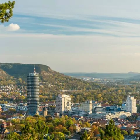 Luftaufnahme von Jena, einer Stadt umgeben von üppigem Grün und Hügeln. Hohe Gebäude, darunter ein markanter Turm, prägen die Landschaft. Der Himmel ist klar mit ein paar Wolkenstreifen. Bäume im Vordergrund rahmen die Stadtszene ein.
