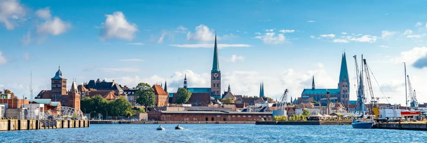 Panoramablick auf die Küstenstadt Lübeck, Deutschland, mit einer Ansammlung historischer Gebäude und Kirchtürme unter einem blauen Himmel mit vereinzelten Wolken. Im Vordergrund liegen Boote und Yachten vor Anker, und das Wasser ist ruhig und spiegelt die malerische Skyline wider – ein perfekter Ort für den Tourismus.