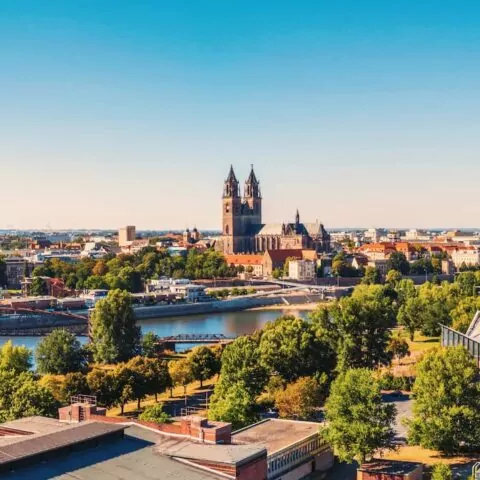 Ein Panoramablick auf die Stadtlandschaft Magdeburgs mit einem Fluss, üppigen grünen Bäumen und historischen Gebäuden. Die Skyline wird vom großen Dom mit seinen zwei Türmen dominiert, während im Hintergrund moderne Gebäude unter einem klaren, blauen Himmel stehen. Perfekt für jeden Reiseführer oder Tourismusliebhaber.