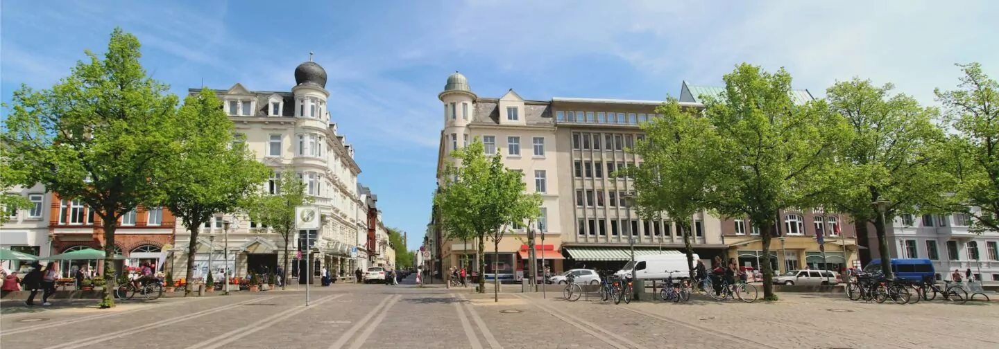 Eine breite Straße, gesäumt von grünen Bäumen, Straßencafés und verschiedenen Gebäuden in unterschiedlichen Architekturstilen, prägt die charmante Promenade von Neumünster. Menschen sitzen in den Cafés, während ein paar Fahrräder und Autos entlang der Straße unter einem klaren blauen Himmel parken.