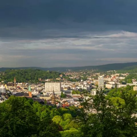 Panoramablick auf die Wuppertaler Stadtlandschaft mit einer Mischung aus modernen und traditionellen Gebäuden, umgeben von üppigen grünen Wäldern und Bäumen. Der Himmel darüber ist teilweise bewölkt, das Sonnenlicht bricht durch und wirft einen warmen Schein über die Stadt. In der Ferne erstrecken sich Hügel.