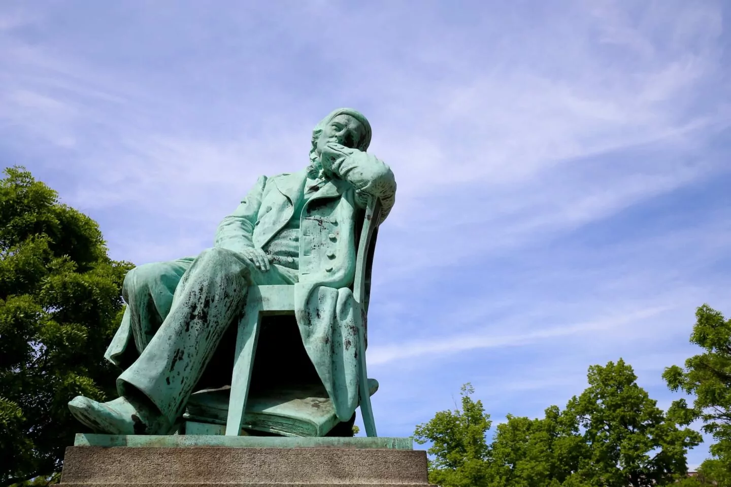 Eine Bronzestatue eines sitzenden Mannes in Kleidung aus dem 19. Jahrhundert, der seinen Kopf auf seine Hand stützt, blickt nachdenklich in die Ferne. Vor einem klaren blauen Himmel mit teilweise sichtbaren grünen Bäumen um den Sockel herum fängt diese Statue in Zwickau einen Moment der Selbstbesinnung ein.