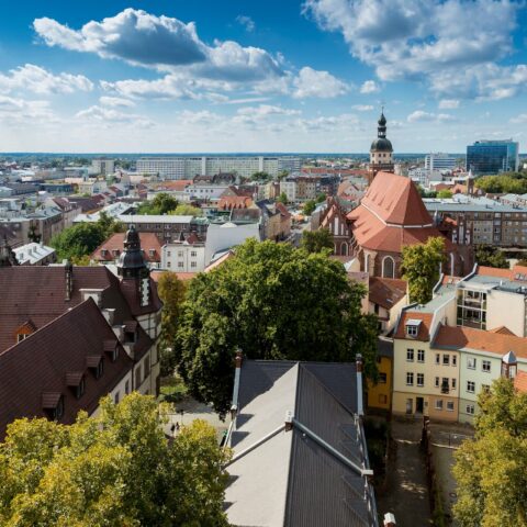 Ein Blick von oben auf die Stadt zeigt eine Mischung aus modernen und traditionellen Gebäuden unter einem klaren blauen Himmel mit vereinzelten Wolken. Inmitten dieses Cottbus-Panoramas sind zwischen den Gebäuden eine markante Kirche mit rotem Dach und verschiedene grüne Bäume zu sehen.