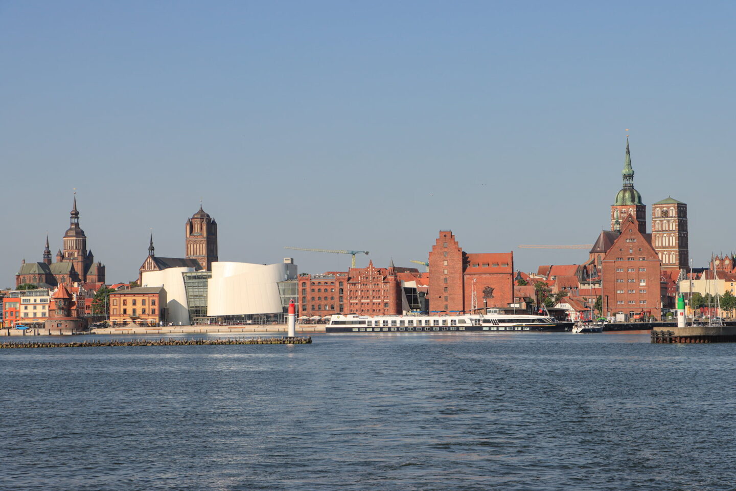 Ein malerischer Blick auf die Uferpromenade der historischen Stadt Stralsund mit markanten Backsteingebäuden und roten Dächern, Kirchtürmen und einem weißen modernen Gebäude. Ein Boot liegt am Ufer und im Vordergrund erstreckt sich das ruhige Wasser unter einem klaren blauen Himmel.