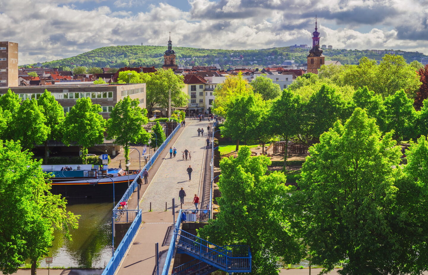 Malerischer Stadtpark in Saarbrücken mit üppigen grünen Bäumen, einem Kanal mit Booten auf der linken Seite und einer Fußgängerbrücke, die zu einer belebten Straße führt. Menschen gehen auf den Wegen im Mittelgrund spazieren. Im Hintergrund sind zwei Uhrtürme und eine Hügellandschaft unter einem teilweise bewölkten Himmel zu sehen.