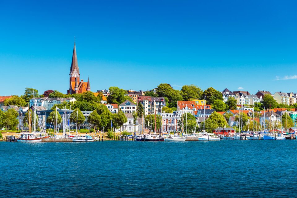Flensburg, eine malerische Küstenstadt, besticht durch farbenfrohe Gebäude, Bäume und eine markante Kirche mit einem hohen Turm. Der Yachthafen im Vordergrund ist voller verschiedener Segelboote und Yachten, alles vor dem Hintergrund eines strahlend blauen Himmels und ruhigen, klaren Wassers.