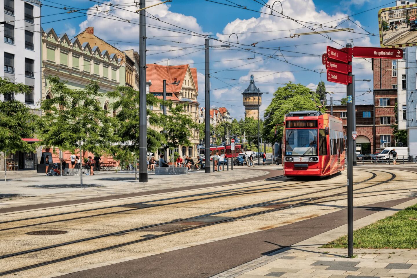 Eine lebendige Stadtszene in Halle mit einer roten Straßenbahn, die auf den Gleisen fährt. Die umliegenden Gebäude sind in verschiedenen Architekturstilen gehalten, darunter ein Turm im Hintergrund. Menschen sitzen und gehen in einer von Bäumen gesäumten Gegend umher und genießen das sonnige Wetter.