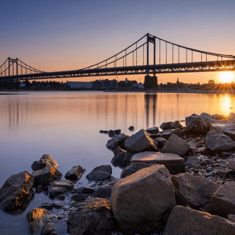Bei Sonnenuntergang überspannt eine Hängebrücke einen ruhigen Fluss in Krefeld und wirft Spiegelbilder auf das Wasser. Der Himmel ist in warmen Orange- und Blautönen gehalten. Im Vordergrund säumen große Felsen das Ufer und verleihen der ruhigen Landschaft Struktur.