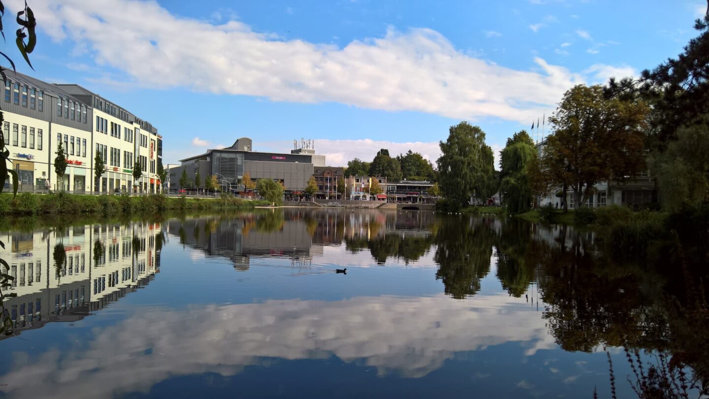 Ein ruhiger See mit Spiegelungen der umliegenden Gebäude und Grünflächen unter einem strahlend blauen Himmel mit vereinzelten Wolken in Neumünster. Eine einsame Ente schwimmt in der Nähe der Mitte und trägt zur friedlichen Atmosphäre bei. Die modernen Gebäude und üppigen Bäume sorgen für ein Gleichgewicht zwischen städtischen und natürlichen Elementen.