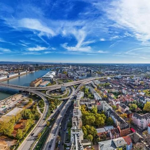 Ein Panoramablick auf eine weitläufige Stadtlandschaft mit einem Fluss, der durch das Zentrum fließt und Brücken freigibt, die verschiedene Teile der Stadt verbinden. Home4Time-Spots wie Handwerkerunterkunft und Monteurwohnung sind zwischen Gebäuden, Grünflächen und Straßen unter einem teilweise bewölkten blauen Himmel mit dünnen Wolken darüber zu sehen.