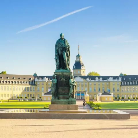 Panoramablick auf das prächtige gelbe Karlsruher Schloss mit einem großen, gepflegten Garten im Vordergrund. In der Mitte ragt eine große Statue einer Person auf einem Sockel hervor. Der Himmel ist klar und blau, mit einer einzelnen weißen Wolke am Himmel. Bäume säumen die Gartenwege.