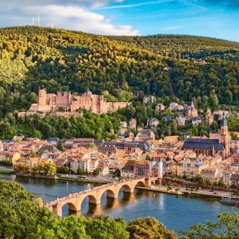 Eine malerische Aussicht auf Heidelberg, Deutschland, mit seinen ikonischen Gebäuden mit roten Dächern, dem stattlichen Heidelberger Schloss auf einem Hügel und der bezaubernden Alten Brücke über den Neckar. Umgeben von üppigem Grün und unter einem strahlend blauen Himmel fängt diese Szene Heidelbergs zeitlose Schönheit ein.