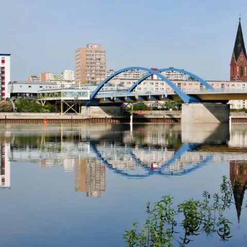 Eine Stadtlandschaft mit einer blau gewölbten Brücke über der ruhigen Oder, in der sich Brücke und Gebäude spiegeln. Im Hintergrund sieht man eine Mischung aus modernen Wohnungen und einer Kirche mit zwei Türmen in Frankfurt, Deutschland. Das Flussufer ist von Grünflächen gesäumt, die einen natürlichen Kontrast zur städtischen Umgebung bilden.