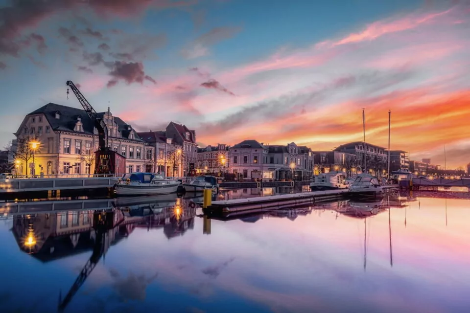 Eine ruhige Hafenszene bei Sonnenuntergang in Oldenburg mit angedockten Booten und stillem Wasser, in dem sich der bunte Himmel spiegelt. Historische Gebäude säumen die Uferpromenade unter einem dramatischen Himmel in Rosa-, Orange- und Blautönen. Ein Kran und Straßenlaternen runden die malerische Aussicht ab.