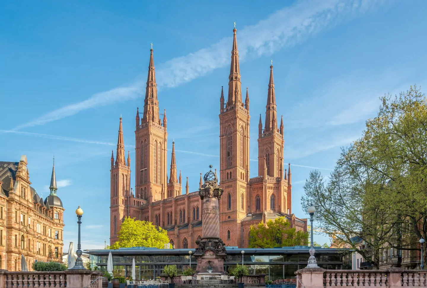 Das Bild zeigt die beeindruckende Fassade der Marktkirche, einer neugotischen Kirche aus rotem Backstein in Wiesbaden, mit fünf hohen Türmen. Im Vordergrund ist ein detailreiches Steindenkmal zu sehen, umgeben von Bäumen und einem klaren blauen Himmel darüber, der die Essenz der malerischen Landschaft Wiesbadens einfängt.