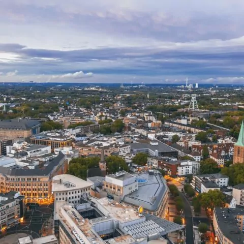 Luftaufnahme der Stadtlandschaft von Bochum in der Abenddämmerung, mit einer Mischung aus moderner und historischer Architektur, Grünflächen mit Bäumen und Straßen mit wenig Verkehr. Der Himmel ist mit Streifen blauer und grauer Wolken bedeckt.