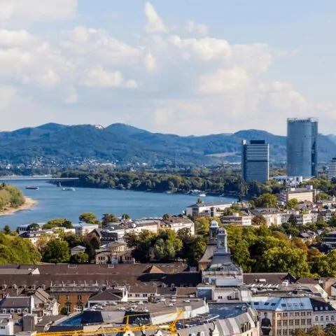 Luftaufnahme von Bonn mit einem durch die Stadt fließenden Fluss, umgeben von Gebäuden, grünen Bäumen und sanften Hügeln im Hintergrund. Moderne Wolkenkratzer und historische Kirchtürme sind sichtbar, mit einer Mischung aus städtischen und natürlichen Landschaften unter einem teilweise bewölkten Himmel.