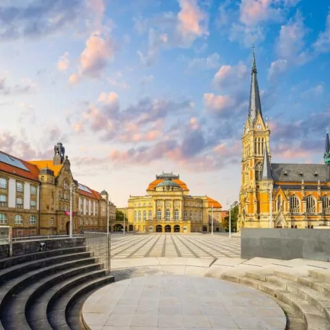 Ein malerischer europäischer Stadtplatz bei Sonnenuntergang in Chemnitz, mit einer großen Kirche mit einem hohen Turm auf der rechten Seite, historischen Gebäuden mit orangefarbenen Dächern und kunstvollen Fassaden auf der linken Seite und Treppen im Vordergrund. Der Himmel ist mit flauschigen rosa Wolken gefüllt.