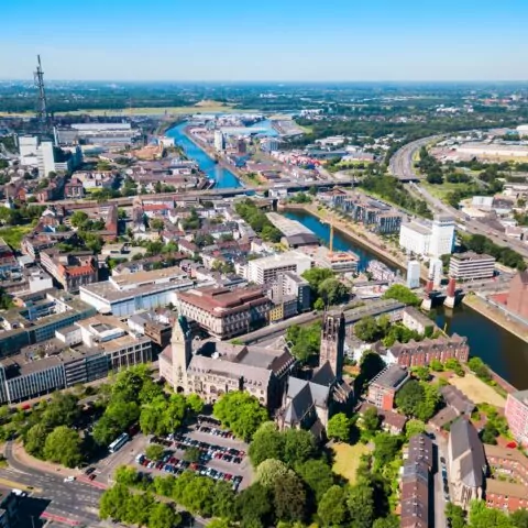 Luftaufnahme von Duisburg, die eine Mischung aus modernen Gebäuden und historischer Architektur zeigt. Im Vordergrund ist eine markante Kirche von Grün umgeben, durch die Mitte verlaufen ein Fluss und mehrere Brücken. Straßen und Infrastruktur sind im gesamten Gebiet sichtbar.