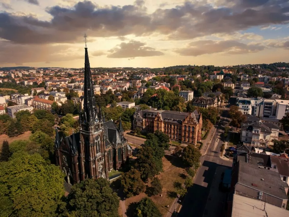 Luftaufnahme einer gotischen Kirche mit hohem Turm in Gera, umgeben von üppigen grünen Bäumen in einer Stadtlandschaft bei Sonnenuntergang. Die Skyline umfasst eine Mischung aus historischen und modernen Gebäuden unter einem teilweise bewölkten Himmel.